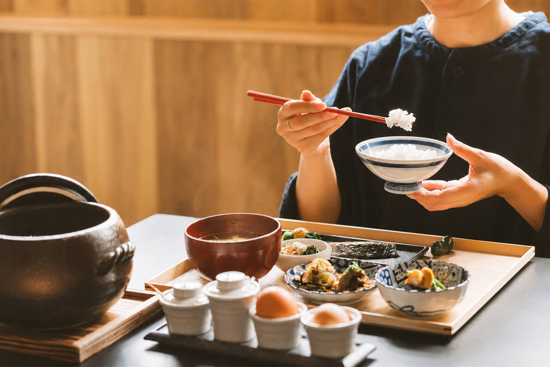 土鍋ごはんの茶碗を手に、味噌汁や小鉢の並ぶ和朝食膳を楽しむ様子